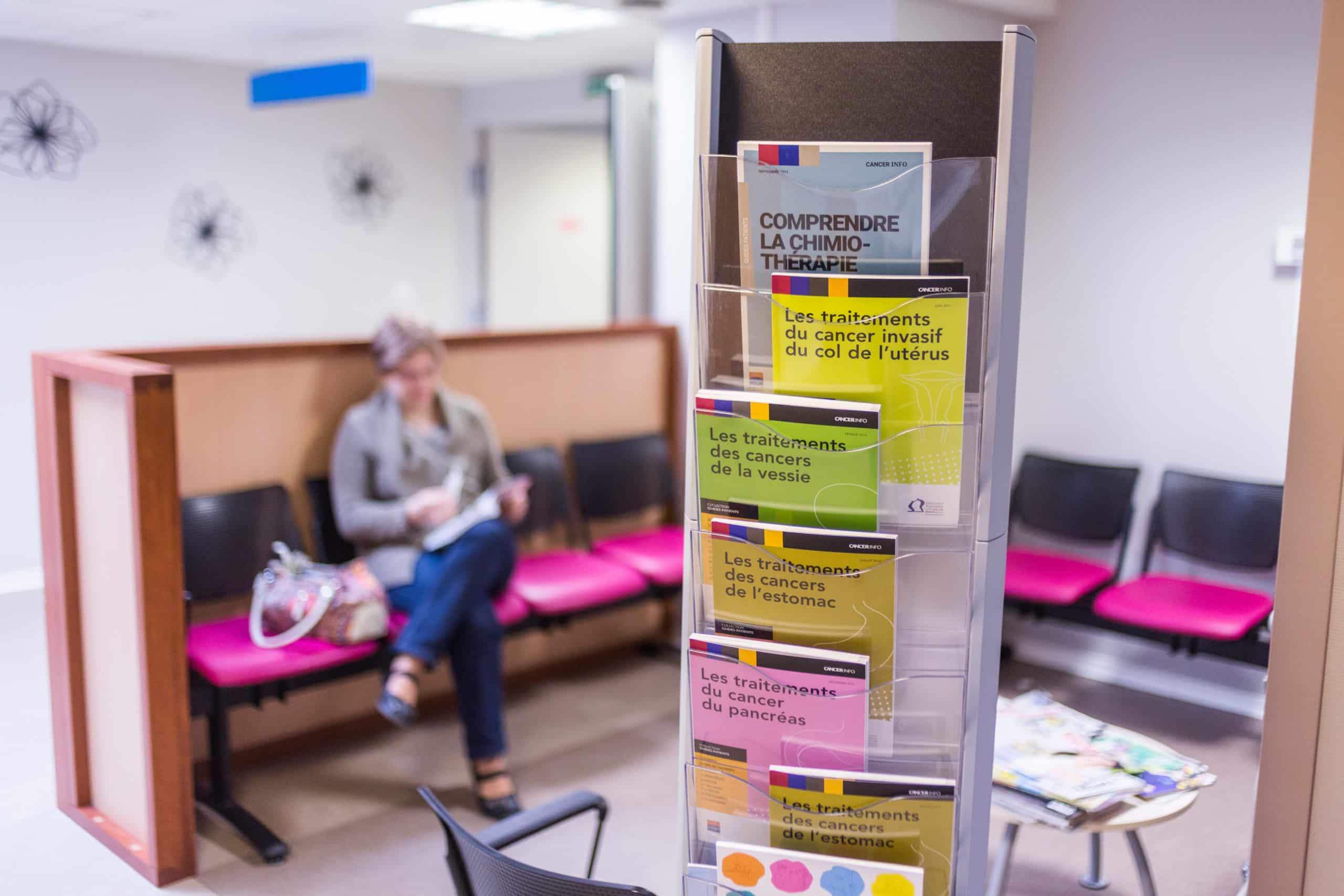 Présentoir de brochures d’information sur les traitements contre le cancer dans une salle d’attente, avec une patiente en arrière-plan lisant un document.